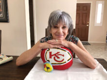 Sun Lakes resident Diane Haver has turned a pandemic hobby into a flourishing part-time job, painting logos of professional and college sports teams on stones from her backyard. Here she is shown with two of her specialties: a hummingbird and a logo of her favorite team, the Kansas City Chiefs. (Photo by Gary Vacin)